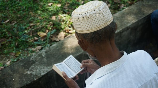 Seorang peziarah membaca Alquran di kuburan massal korban tsunami di Ulee Lheue, Banda Aceh, Kamis (26/12/2024). [Suara.com/Habil Razali]