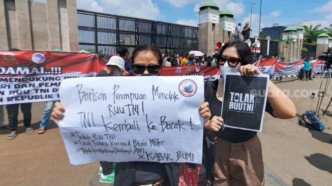 Dua wanita membawa poster berisikan penolakan terhadap RUU TNI berdiri di depan gerbang Gedung DPR/MPR, Senayan, Jakarta. (Suara.com/Novian)