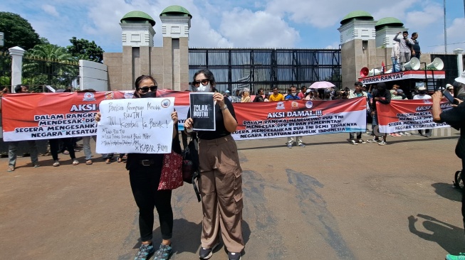 Dua wanita tetap berdiri tegap membawa poster berisikan penolakan terhadap RUU TNI di depan gerbang Gedung DPR/MPR, Senayan, Jakarta. (Suara.com/Novian)