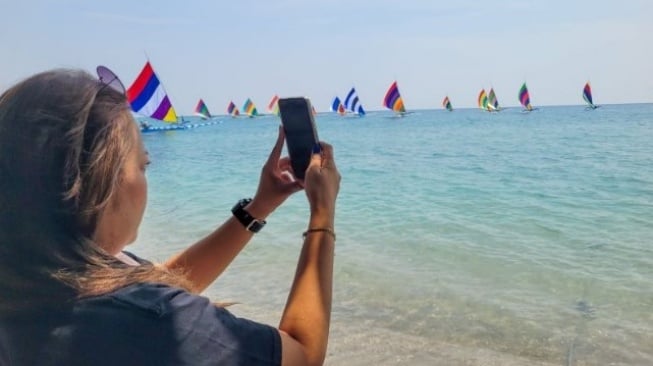 Seorang pengunjung mengabadikan gambar kegiatan lomba balap perahu layar di kawasan Wisata Bahari Pasir Putih Situbondo, jawa Timur, Minggu (24/8/2025) [Suara.com/ANTARA]