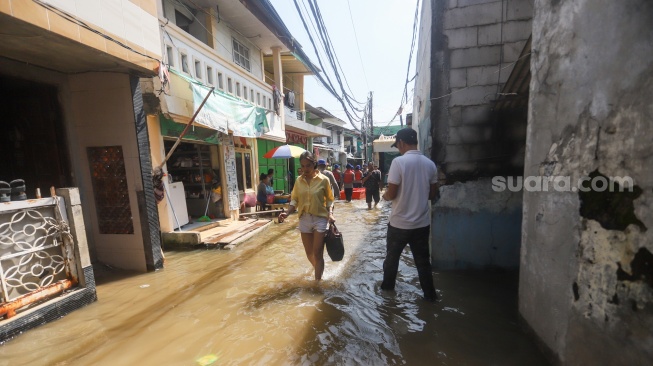 Pengguna jalan melintasi banjir rob di Muara Angke, Jakarta, Jumat (5/12/2025). [Suara.com/Alfian Winanto]
