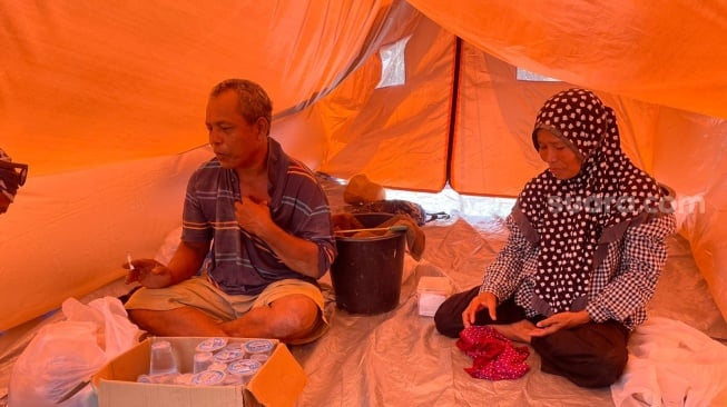 Ali Rasyid&mdash;atau yang lebih dikenal warga dengan sapaan Wujud (kiri) bersama istri Salma (kanan), di tenda kecil di Gampong Sukajadi. [Suara.com/Iskandar]