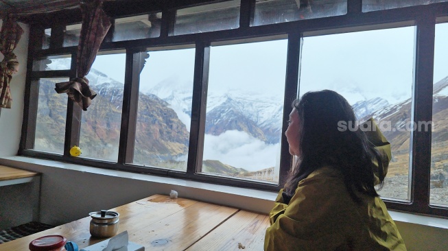 Seorang pendaki wanita menatap pemandangan dinding gunung bersalju dan gumpalan awan dari balik jendela sebuah penginapan di kawasan Himalaya, Nepal, Jumat (29/5/2026). [Suara.com/Rizka Chaerani] 