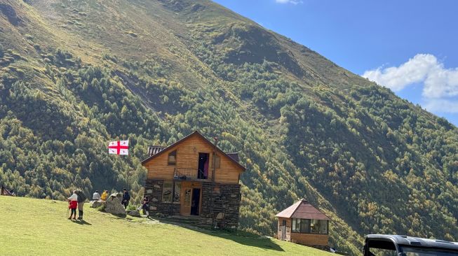 Wisatawan berada di sekitar bangunan kayu dengan latar perbukitan Pegunungan Kaukasus di wilayah Svaneti, Georgia, Minggu (27/9/2026). [Suara.com/Rizka Chaerani]