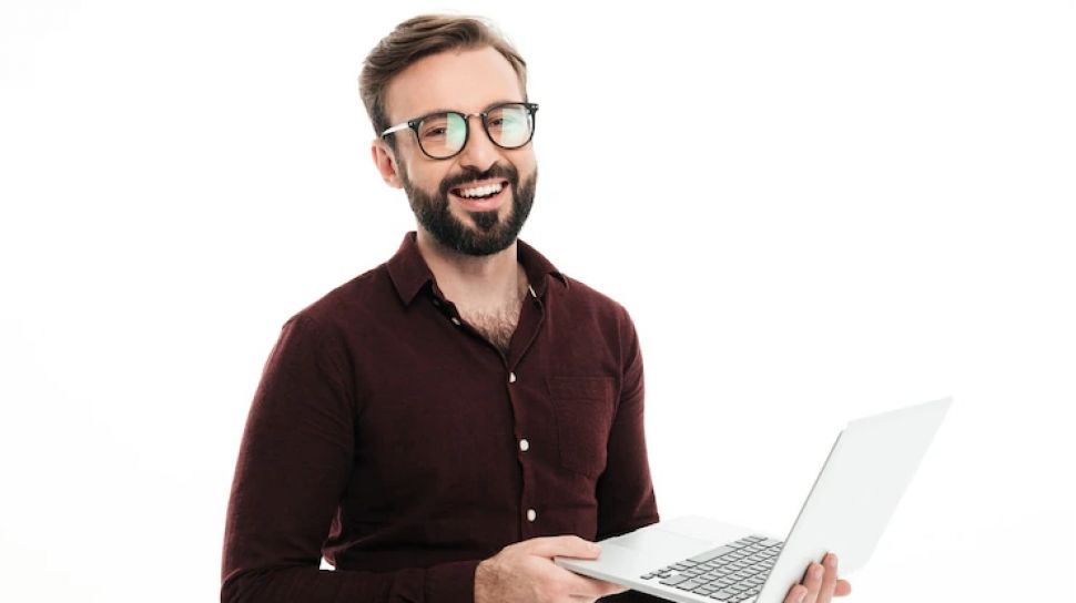  A man wearing glasses is smiling while holding an open laptop in front of a white background.