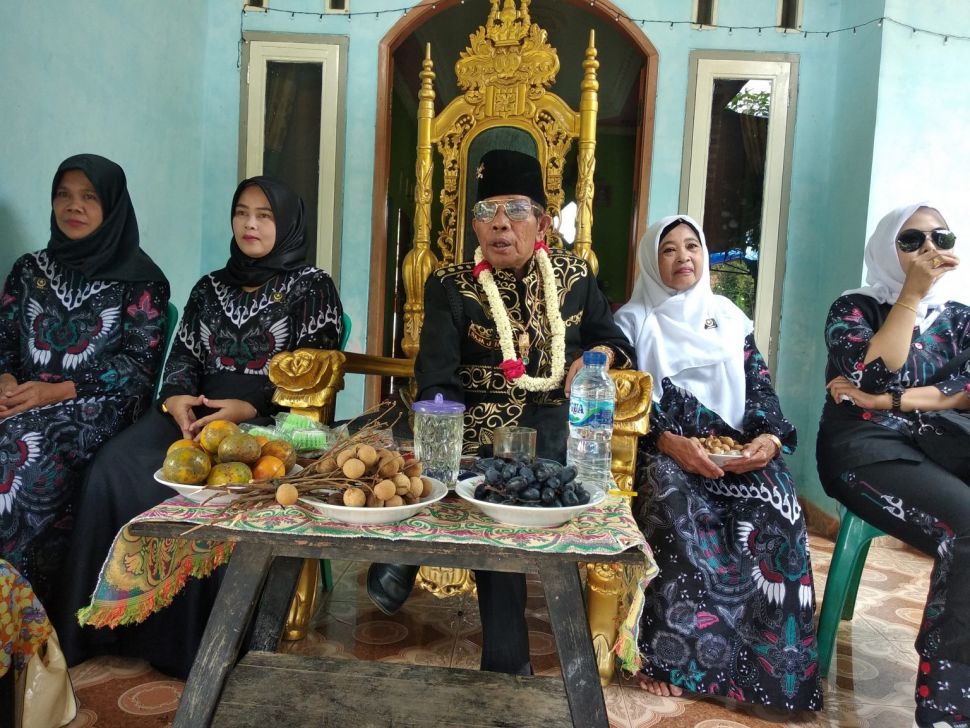 Iskandar Jamaludin Firdos pria yang dipanggil raja oleh warga duduk di kursi singgasana ditemani empat perempuan saat serah terima kunci rumah kepada warga. Ia membangun 30 rumah warga miskin di Pandeglang. [BantenHits]