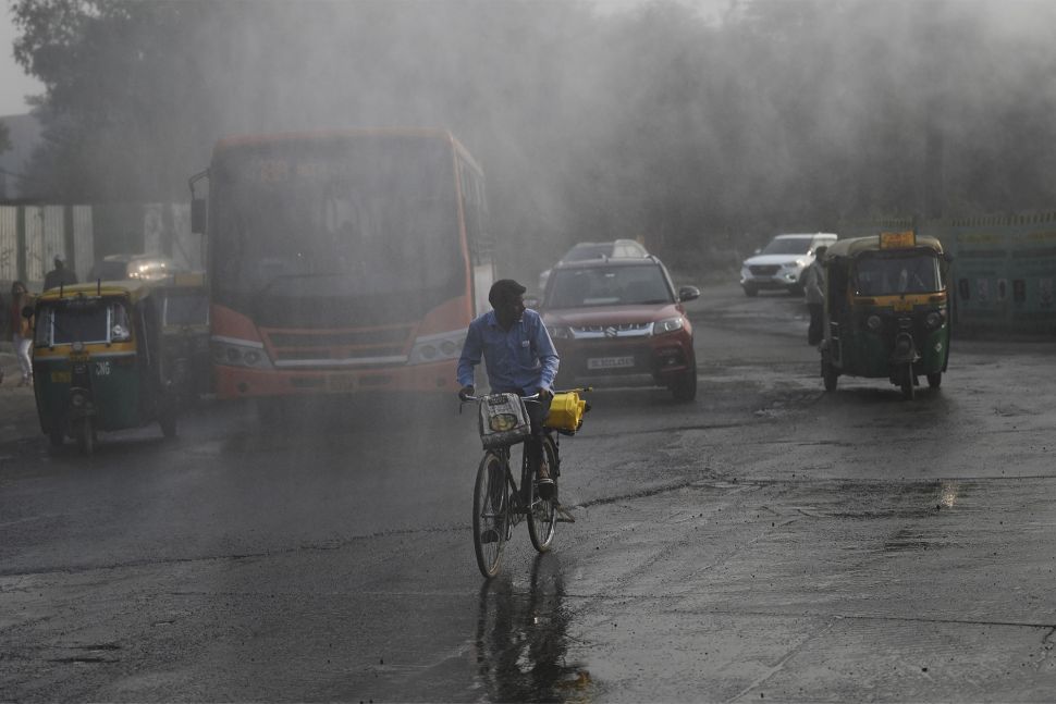 Para komuter melintas melewati alat anti-kabut asap yang menyemprotkan air untuk mengekang polusi udara di New Delhi, India, pada (16/11/2021). [MONEY SHARMA / AFP]