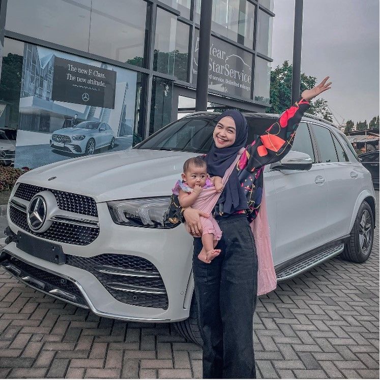 Ria Ricis menggendong baby Moana di depan sebuah mobil Mercedes-Benz berwarna putih. Diketahui ia membeli mobil tersebut untuk anaknya. [Instagram/@riaricis1795]